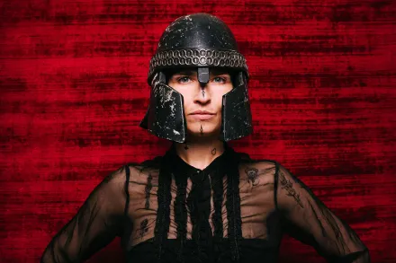 Frau schwarzgekleidet mit Ritter*innenhelm auf dem Kopf vor schwarz-rotem Hintergrund