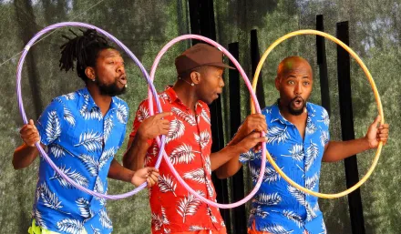 Drei Männer in Hawaiihemden mit Palmenmuster, zwei in Rot, einer in Blau, stehen nebeneinander und halten sich Hula Hoop-Reifen vor den Körper, schauen durch die Reifen