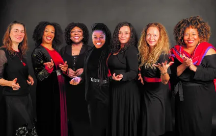 7 Frauen vor grauem Hintergrund. Die Frauen sind festlich in schwarz-rot gekleidet und lächeln und lachen freundlich in die Kamera