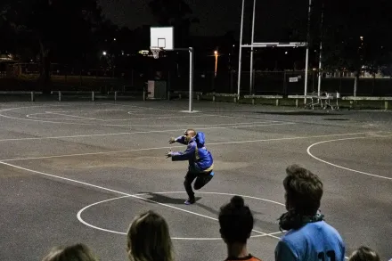 Eine Person auf einem Sportplatz, in einer dynamischen Bewegung, im Vordergrund ist Publikum zu sehen. Die Szene spielt im Freien bei Dunkelheit