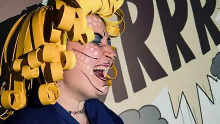 Eine kräftig geschminkte Person mit gelber Perücke, von der Seite aus fotografiert, die mit weit geöffnetem Mund und geschlossenen Augen singt/schreit. Im Hintergrund eine Comic-Zeichnung mit dunklen Wolken und "RRRR" in einem gelben Feld mit Zacken