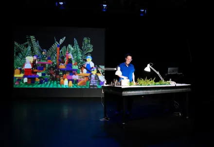 Eine Frau präsentiert auf der Bühne eine LEGO-Szene, die groß im Hintergrund projiziert wird.