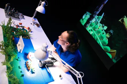 Person baut eine Szene mit LEGO-Steinen auf einem Tisch, daneben Pflanzen und zwei weiße Schreibtischlampen, im Hintergrund Projektion einer ähnlichen LEGO-Szene
