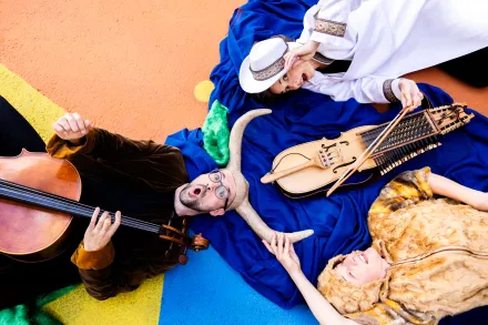 Drei Personen liegen auf buntem Boden, eine spielt Cello, eine trägt weißen Hut und hält eine Nyckelharpa, eine trägt Fellumhang und hält ein Horn