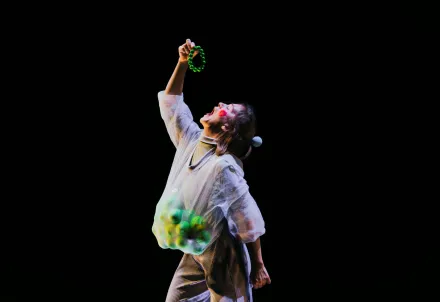 Person in weißem Kostüm mit Netzstoff und grünen Bällen hebt einen grünen Ring in die Luft und schaut mit offenem Mund nach oben, vor schwarzem Hintergrund.