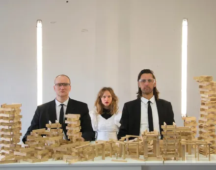 Drei Personen sitzen nebeneinander an einem Tisch, der mit zahlreichen Holzbausteinen bedeckt ist. Die Personen tragen dunkle Anzüge mit weißen Hemden, die mittlere Person ein weißes Kleid mit Puffärmeln. Im Hintergrund sind zwei vertikale Lichtstreifen zu sehen. Die Holzbausteine sind zu verschiedenen, teils instabil wirkenden Türmen und Konstruktionen gestapelt.