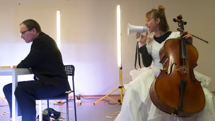 Im Vordergrund sitzt eine Person in einem dunklen Anzug an einem weißen Tisch und blickt abgewandt. Daneben sitzt eine weitere Person in einem weißen, voluminösen Kleid auf einem Stuhl und hält ein Cello sowie ein Megafon. Im Hintergrund stehen zwei senkrechte Lichtstäbe vor einer hellen Wand, auf dem Boden liegen verstreute Holzstücke.