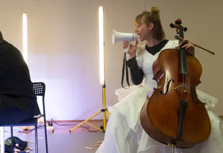 Im Vordergrund sitzt eine Person in einem dunklen Anzug an einem weißen Tisch und blickt abgewandt. Daneben sitzt eine weitere Person in einem weißen, voluminösen Kleid auf einem Stuhl und hält ein Cello sowie ein Megafon. Im Hintergrund stehen zwei senkrechte Lichtstäbe vor einer hellen Wand, auf dem Boden liegen verstreute Holzstücke.