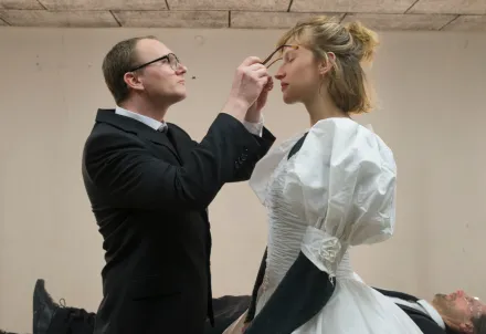 Eine Frau in einem weißen Kleid mit voluminösen Ärmeln steht seitlich im Vordergrund. Ein Mann im dunklen Anzug setzt ihr eine Brille auf. Im Hintergrund liegt eine weitere Person in dunkler Kleidung auf einem Tisch. Zwei vertikale Lichtstäbe stehen links und rechts am Rand des Raumes.
