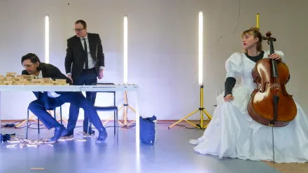 Im Vordergrund sitzt eine Person in einem Hochzeitskleid auf einem Stuhl und hält ein Cello. Rechts und links hinter ihr stehen senkrechte Lichtstäbe auf Stativen. Links im Bild sitzen und stehen zwei Personen in dunklen Anzügen an einem weißen Tisch, auf dem Holzklötze verteilt sind; einige Klötze liegen auf dem Boden.