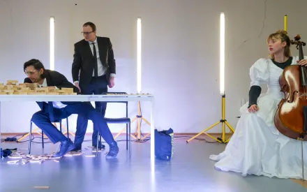 Im Vordergrund sitzt eine Person in einem Hochzeitskleid auf einem Stuhl und hält ein Cello. Rechts und links hinter ihr stehen senkrechte Lichtstäbe auf Stativen. Links im Bild sitzen und stehen zwei Personen in dunklen Anzügen an einem weißen Tisch, auf dem Holzklötze verteilt sind; einige Klötze liegen auf dem Boden.