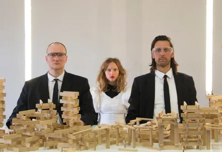 Drei Personen sitzen nebeneinander an einem Tisch, der mit zahlreichen Holzbausteinen bedeckt ist. Die Personen tragen dunkle Anzüge mit weißen Hemden, die mittlere Person ein weißes Kleid mit Puffärmeln. Im Hintergrund sind zwei vertikale Lichtstreifen zu sehen. Die Holzbausteine sind zu verschiedenen, teils instabil wirkenden Türmen und Konstruktionen gestapelt.