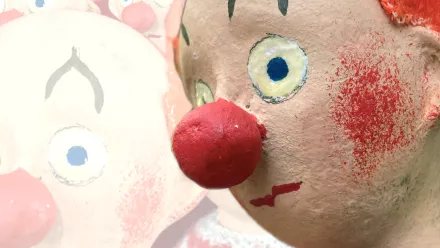 Deteilaufnahme des Gesichts einer Clown-Puppe mit roter Nase, roten Wangen und orangen Haaren. Im Hintergrund dasselbe Foto, aber ganz hell bzw transparent