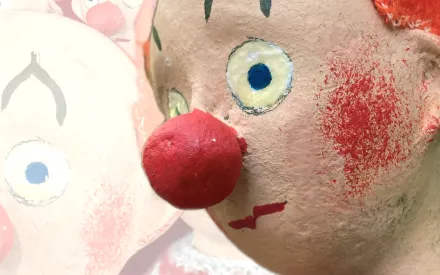 Deteilaufnahme des Gesichts einer Clown-Puppe mit roter Nase, roten Wangen und orangen Haaren. Im Hintergrund dasselbe Foto, aber ganz hell bzw transparent