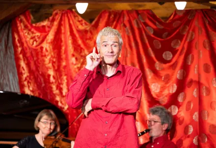 Ein Künstler in rotem Hemd steht vor einem roten Vorhang und spricht auf der Bühne.