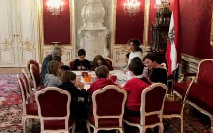 Eine Gruppe von Kindern sitzt mit dem österreichischen Bundespräsidenten Alexander van der Bellen und seiner Frau, Doris Schmidauer,um einen Tisch in der österreichischen Präsidentschaftskanzlei, einem prunkvollen Raum mit roten Wänden und goldenen Verzierungen. Die Stühle sind mit rotem Stoff bezogen und haben weiße Holzrahmen. Auf dem Tisch liegen verschiedene Bastelmaterialien. Im Hintergrund stehen ein großer, weißer Kachelofen und eine österreichische Fahne. Der Boden ist mit einem gemusterten Teppich bedeckt.