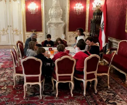 Eine Gruppe von Kindern sitzt mit dem österreichischen Bundespräsidenten Alexander van der Bellen und seiner Frau, Doris Schmidauer,um einen Tisch in der österreichischen Präsidentschaftskanzlei, einem prunkvollen Raum mit roten Wänden und goldenen Verzierungen. Die Stühle sind mit rotem Stoff bezogen und haben weiße Holzrahmen. Auf dem Tisch liegen verschiedene Bastelmaterialien. Im Hintergrund stehen ein großer, weißer Kachelofen und eine österreichische Fahne. Der Boden ist mit einem gemusterten Teppich bedeckt.