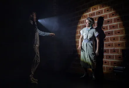 Zwei Personen in einer dunklen Szene: Eine leuchtet mit einer Taschenlampe auf die andere, die erschrocken an einer Backsteinwand steht.