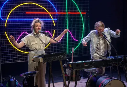 Zwei Personen stehen vor bunten Neonlinien an Keyboards und Mikrofonen; sie performen mit energiegeladener Gestik und musikalischem Ausdruck auf der Bühne.