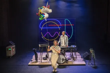 Zwei Musiker auf einer Bühne mit Schlagzeug und Synthesizern, im Hintergrund bunte Neonlichter und ein Einhorn-Luftballon