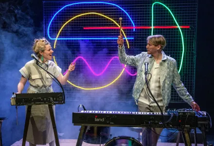 Zwei Personen stehen an Keyboards auf der Bühne, umgeben von bunten Neonlinien im Hintergrund, und zeigen mit erhobenen Schlägeln spielerisch aufeinander.