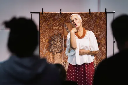 Frau in weißer Bluse und rotem Rock steht vor einem Wandteppich mit floralem Muster und spricht vor Publikum