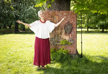 Person mit weißem Haar, weißer Bluse und rotem Rock steht lächelnd mit ausgebreiteten Armen im Park vor einem Baum und einem Wandteppich mit Einhornmotiv.