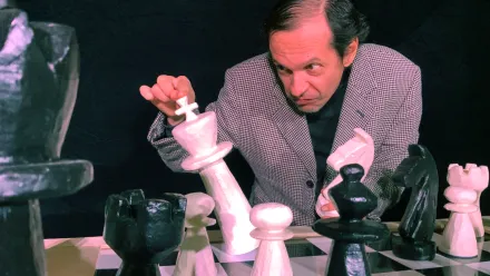 EIn grimmig dreinblickender Mann mit seitlich glattgekämmten Haaren bei einem Schachspiel mit ca 30 Zentimeter hohen Figuren, kippt mit seiner rechten Hand den weißen König.