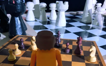 Im Hintergrund ein großes Schachbrett mit Figuren, im Vordergrund ein kleineres Schachbrett mit Figuren, beides aus Holz, davor steht eine Playmobil-Figur, den Schachfeldern zugewandt, de*r*m Betrachter*in den Rücken zugewandt