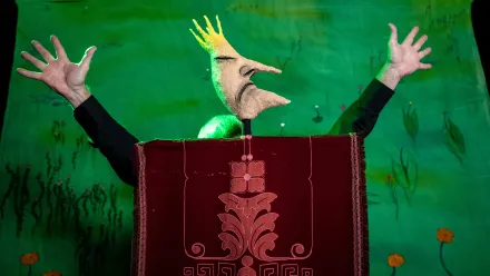 Eine lustige Königsfigur mit langer Nase und spitzem Kinn, beide Arme nach oben gestreckt, die Finger auseinandergespreizt. im Hintergrund eine gemalte Wiese mit orangen Blumen