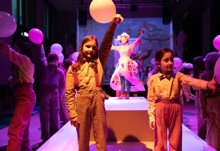 Kinder mit Ballons auf der Bühne, im Vordergrund zwei Kinder mit Hosenträgern, im Hintergrund eine bunt gekleidete Figur mit Blumenkrone.