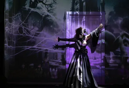 Person in schwarz-weißem Kostüm mit ausdrucksstarkem Make-up, auf der Bühne vor einer Projektion mit Spinnennetzen und dunklem Schloss.