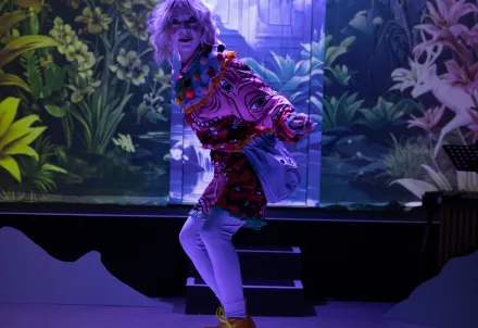 Person mit blondem welligem Haar und buntem Kostüm voller Muster und Pompons, auf der Bühne vor einer tropischen Pflanzenkulisse.