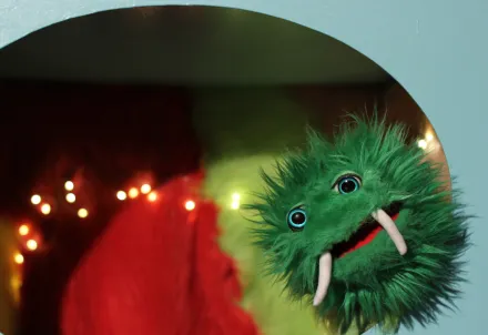 Grüne, pelzige Handpuppe mit großen Augen und zwei langen, weißen Stoßzähnen vor rotem und gelbgrünem Hintergrund mit Lichterkette
