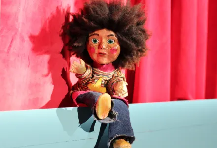 Puppe mit lockigem Haar, Sommersprossen, bunter Kleidung und Jeans sitzt vor rotem Vorhang auf hellblauem Podest
