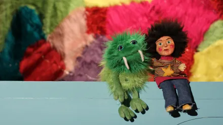 Grüne Plüschfigur mit Stoßzähnen neben Puppe mit dunklem Haar und buntem Pullover vor bunter Wand.