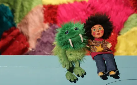 Grüne Plüschfigur mit Stoßzähnen neben Puppe mit dunklem Haar und buntem Pullover vor bunter Wand.