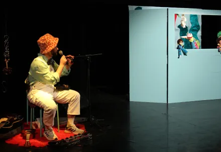 Ein Mann mit orangegemustertem Hut sitzt auf einem Stuhl und singt in ein Mikrofon, gegenüber eine hellblaue Puppenbühne mit mehreren bunten Handpuppen.