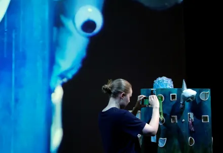 Eine Person filmt eine fantasievolle Miniaturkulisse in Blautönen, während im Hintergrund eine vergrößerte Meeresfigur projiziert wird.