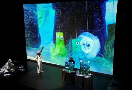 Eine Performerin interagiert auf der Bühne mit einer groß projizierten Unterwasserwelt voller bunter, lächelnder Meeresfiguren.
