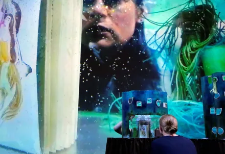 Person betrachtet eine fantasievolle Miniaturbühne vor einer groß projizierten, farbintensiven Unterwasser- oder Traumwelt-Szene.