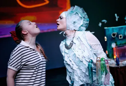 Die Donaunixe hält mehrere kleine Wesen mit Tentakeln in der Hand. Neben ihr steht eine andere Performerin. Sie singen sich entgegen.