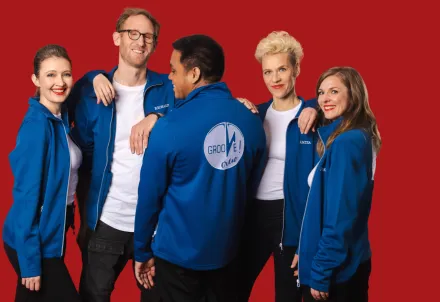 Fünf Personen stehen vor einem einfarbig roten Hintergrund. Sie tragen blaue Jacken, von denen eine auf dem Rücken einen großen, runden Aufdruck mit dem Schriftzug "Groove Crew" Die Personen stehen eng beieinander, einige blicken sich an, andere schauen in verschiedene Richtungen.