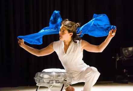 Eine Tänzerin kniet auf der Bühne und hält ein wehendes blaues Tuch über einem Ventilator.