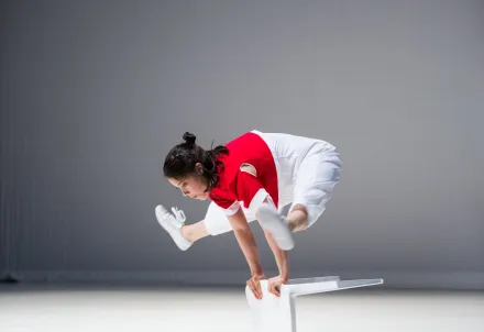 Eine Tänzerin springt mit gestreckten Beinen über einen weißen Stuhl auf der Bühne.