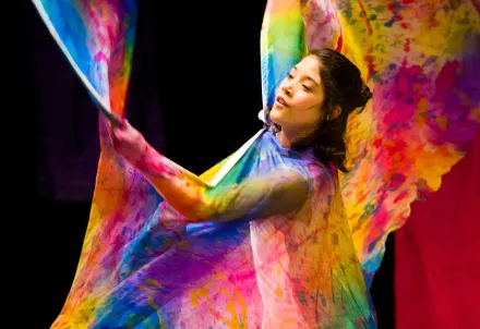 Eine Tänzerin bewegt bunte, wehende Stoffbahnen in leuchtenden Farben auf der Bühne.