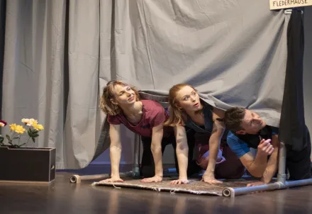 Drei Performer*innen kriechen neugierig unter einem grauen Vorhang hervor.