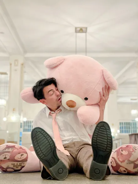 Person sitzt auf dem Boden in einem hellen Raum, hält einen großen rosa Teddybär und trägt weiße Hemd und rosa Krawatte