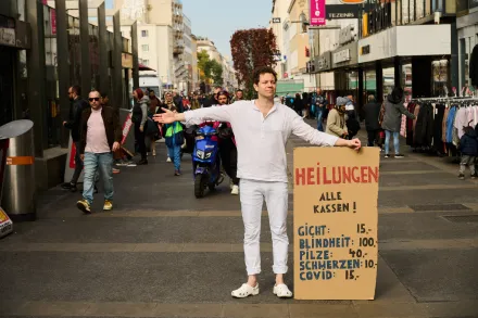 Person in weißer Kleidung steht auf belebter Einkaufsstraße und hält Schild mit Preisen für Heilungen bei verschiedenen Krankheiten