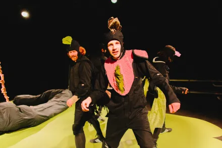 Person in schwarzem Overall mit pinkem, strukturiertem Kragen und grünem Detail auf einer Bühne mit gelbem Boden